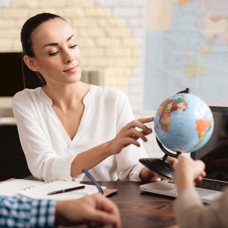 Consulente di agenzia turistica mostra destinazioni sul mappamondo a clienti in ufficio, con quaderno, penna e laptop sul tavolo.