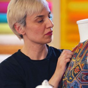 Elisa Pizziol, sarta modellista e docente di sartoria, lavora su un abito colorato su manichino, applicando dettagli di cucito a mano in laboratorio.