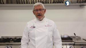 Chef Gaetano Simonato in cucina con giacca bianca personalizzata “Tano Passami l’Olio”, durante una lezione per l’Agenzia Formativa Dante Alighieri.