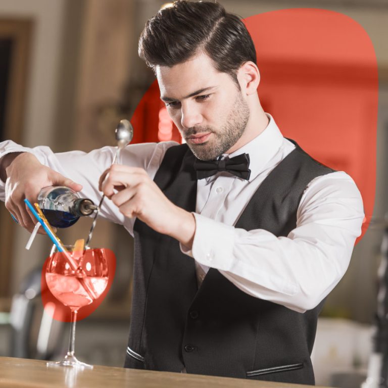 Barman professionista in camicia bianca e gilet nero prepara un cocktail al bancone, versando liquore in un bicchiere con ghiaccio e decorazione.
