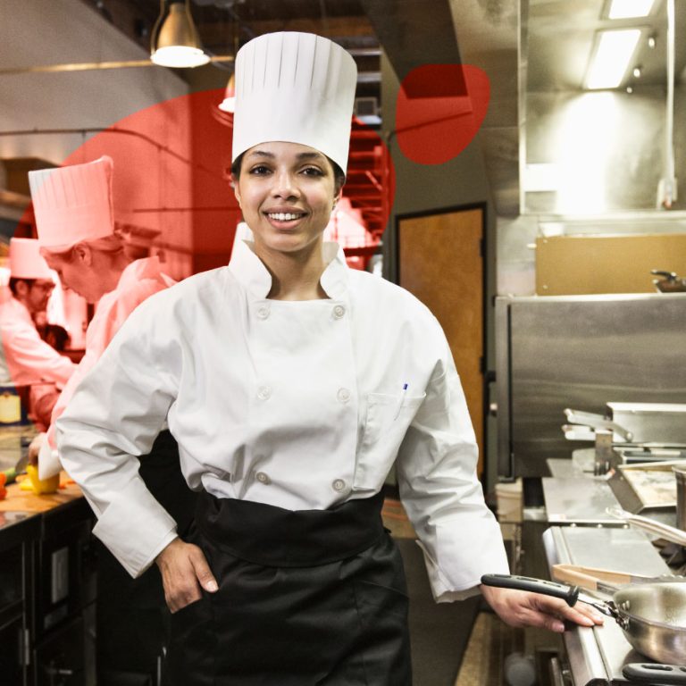 Cuoca professionista in uniforme bianca e toque da chef lavora in cucina industriale, affiancata da colleghi impegnati nella preparazione dei piatti.