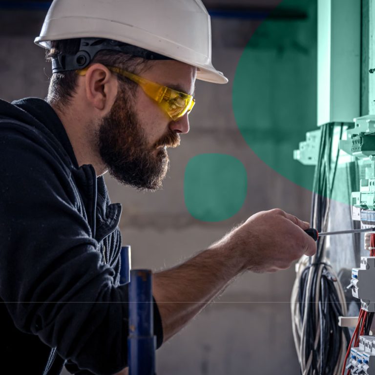 Electricista professional amb casc i ulleres de protecció treballant en un quadre elèctric, utilitzant un tornavís per connectar els circuits de manera segura.