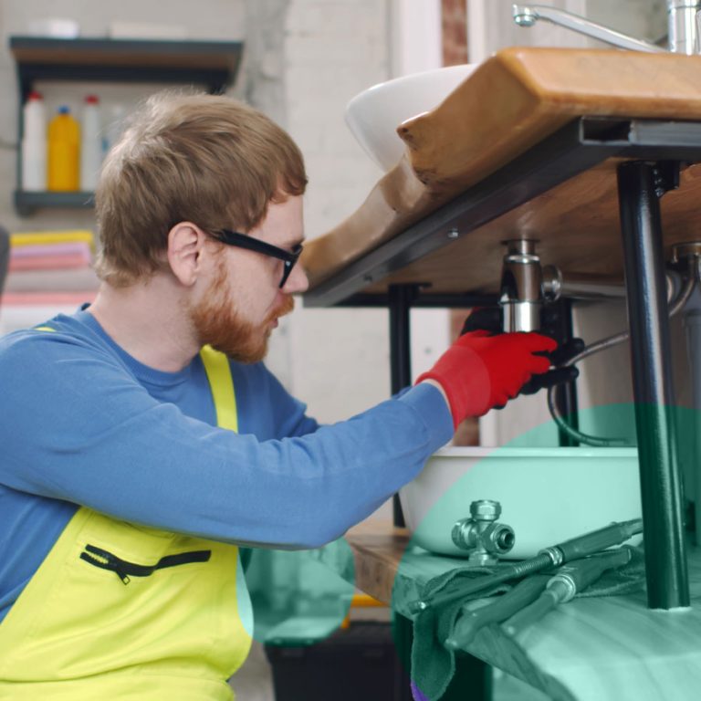 Fontanero profesional repara el desagüe de un lavabo bajo la encimera, con herramientas de trabajo y guantes protectores, durante una intervención técnica.