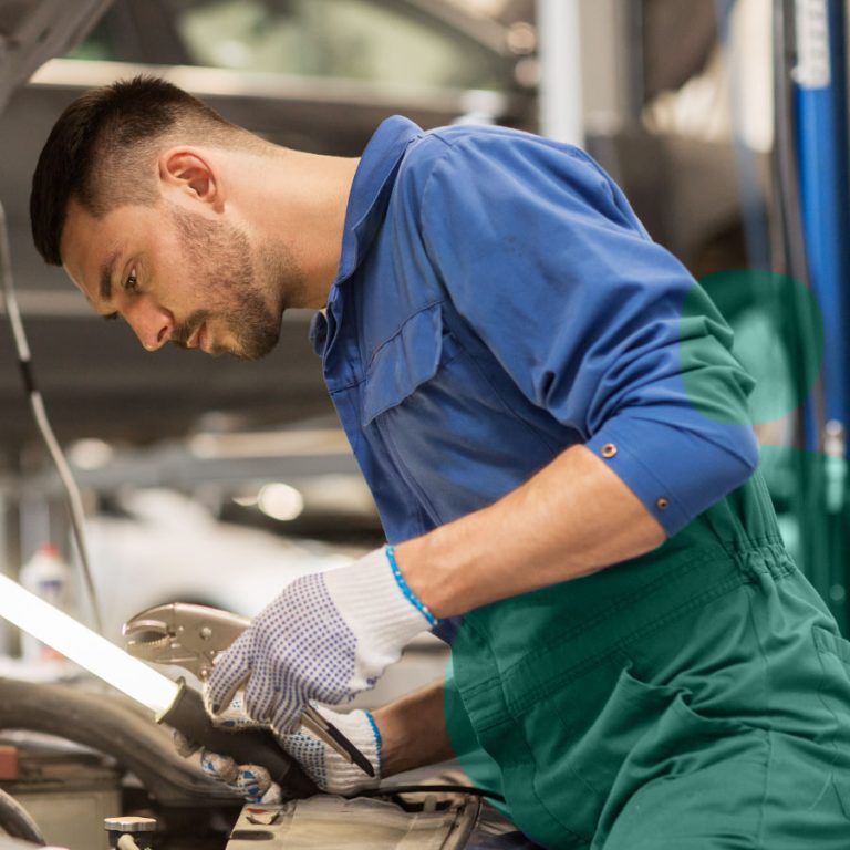 Mecánico en mono de trabajo azul y verde revisa el motor de un coche con lámpara de inspección, llevando guantes protectores en el taller.