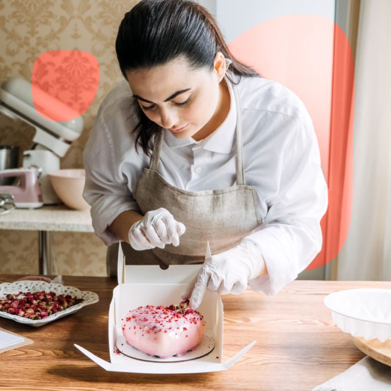 Pasticcera in camice bianco e grembiule decora con cura una torta glassata rosa, aggiungendo petali essiccati in laboratorio di pasticceria artigianale.
