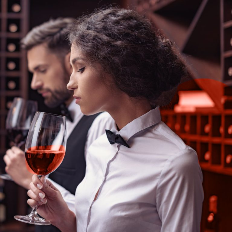 Sommelier in camicia bianca e papillon mentre degusta un calice di vino rosato in una cantina con bottiglie esposte.