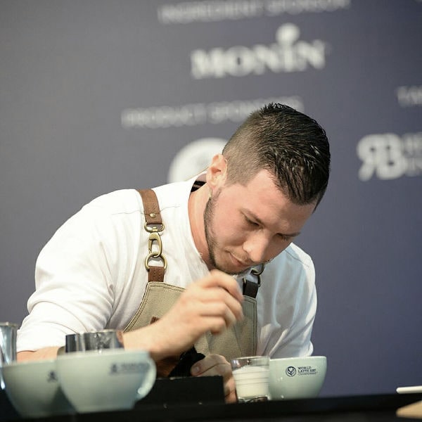 Matteo Beluffi in gara di latte art, con microfono e grembiule, esegue una decorazione su cappuccino.